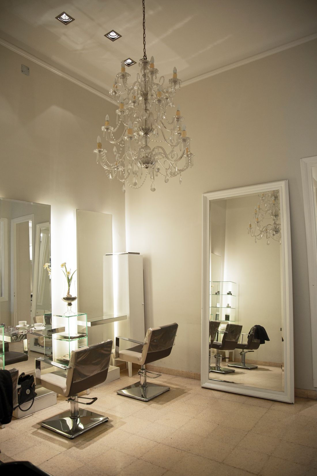 Elegant salon corner with a crystal chandelier, two cream-and-brown styling chairs with chrome bases, a tall white-framed floor mirror, glowing mirror panels and glass shelving, and a vase with a flower.