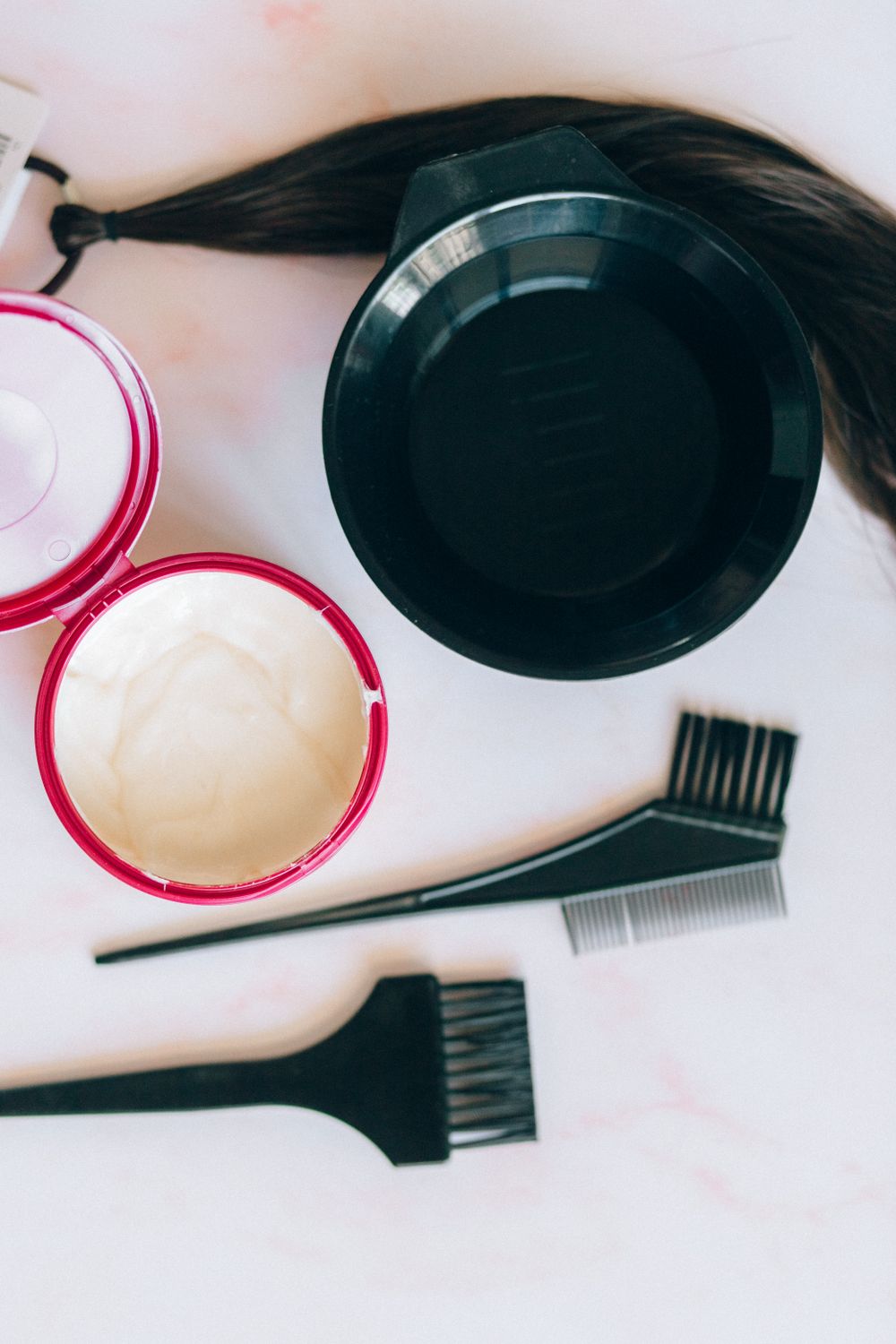 Flat lay on pink-veined marble: a black mixing bowl, a loose tress of dark brown hair tied with a band, an open round tub with a red rim holding cream, a black tint brush and a black comb brush.