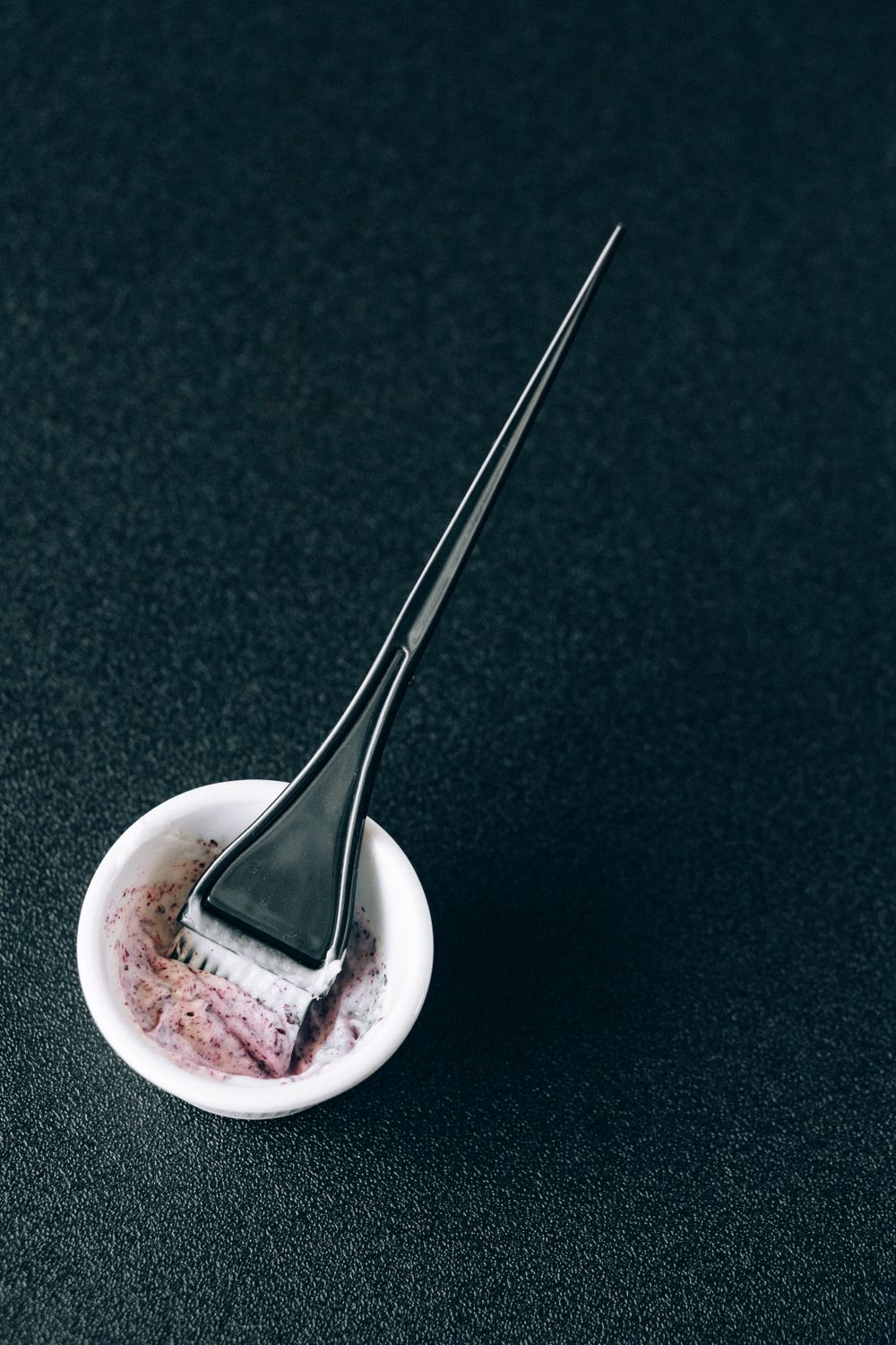 A black tint brush resting in a small white bowl with a purple-and-white colour mixture, on a dark textured surface.