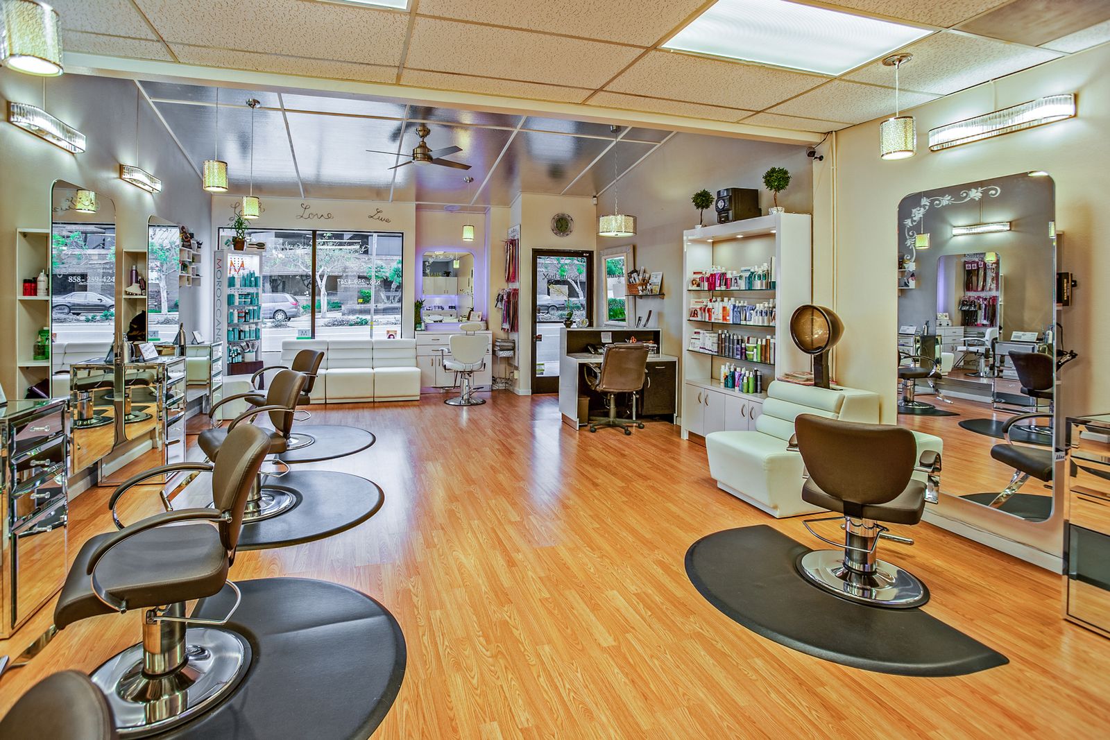 Wide interior of a bright hair salon: wooden floor, several brown styling chairs on black round mats, wall-length mirrors and shelves of hair products, a white leather sofa and a shopfront window.